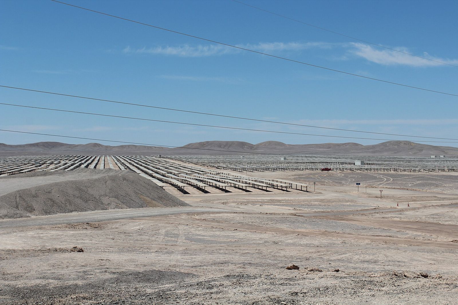 Hileras de paneles solares de la planta fotovoltaica Cerro Dominador en el desierto, con cables de una línea eléctrica cruzando el cielo y cerros al fondo