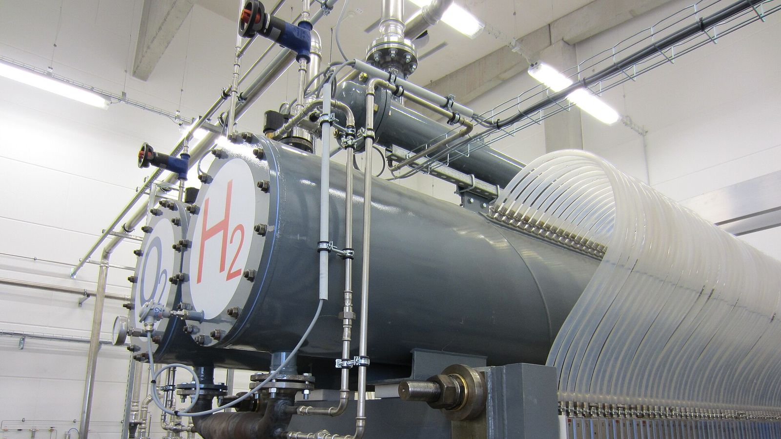 Electrolizador en una sala de la central híbrida de Prenzlau: depósitos cilíndricos grises rotulados H2 (hidrógeno) y O2 (oxígeno), tuberías de acero y un stack de celdas con decenas de mangueras transparentes