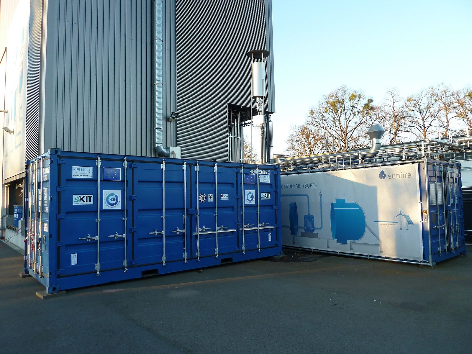 Protótipo power-to-gas Helmeth em dois contêineres ao lado de um edifício industrial: à esquerda, o módulo azul de metanação de CO2; à direita, o de eletrólise de vapor a alta temperatura (SOEC)