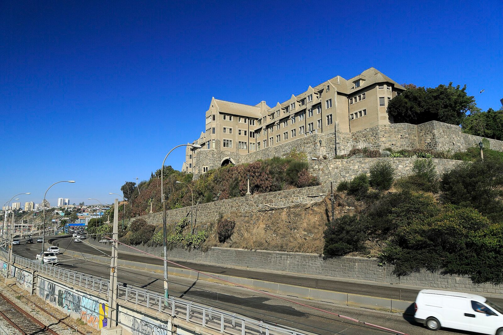 Edifício da Casa Central da Universidad Técnica Federico Santa María em Valparaíso, sobre muros de pedra na encosta, com a avenida España e a linha do trem em primeiro plano